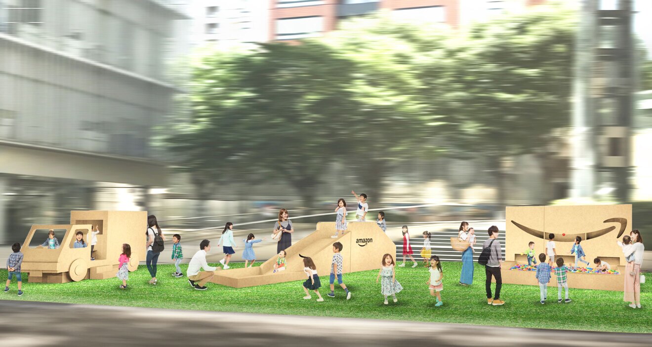 芝生広場に設置された段ボール遊具。子どもが乗って遊べる配送トラック型の遊具と滑り台、ボールプールの3つ。遊具はベージュ色でAmazonのロゴが描かれている。遊ぶ子どもたちと見守る保護者がいる。