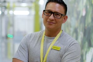 An image of a man smiling for a photo while standing in an Amazon Style store. He is wearing a neon yellow lanyard and a neon yellow name tag. 