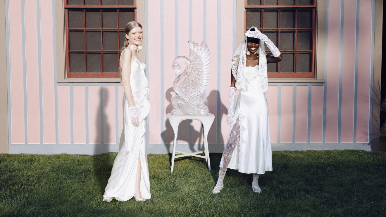 Two women wear wedding gowns and stand outside in front of a pink wall.