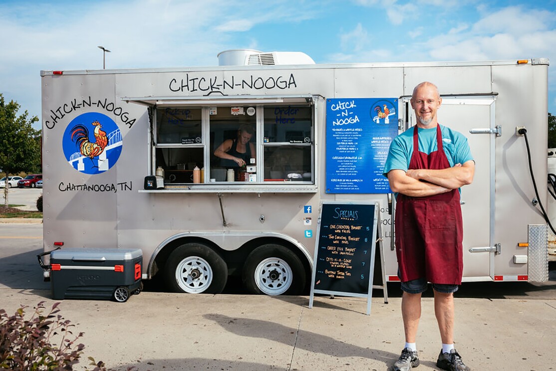 A man wearing a maroon apron stands with his arms crossed, in front of a  silver CHICK-N-NOOGA food truck. A sign displays the specials in neon orange text on a black background. 