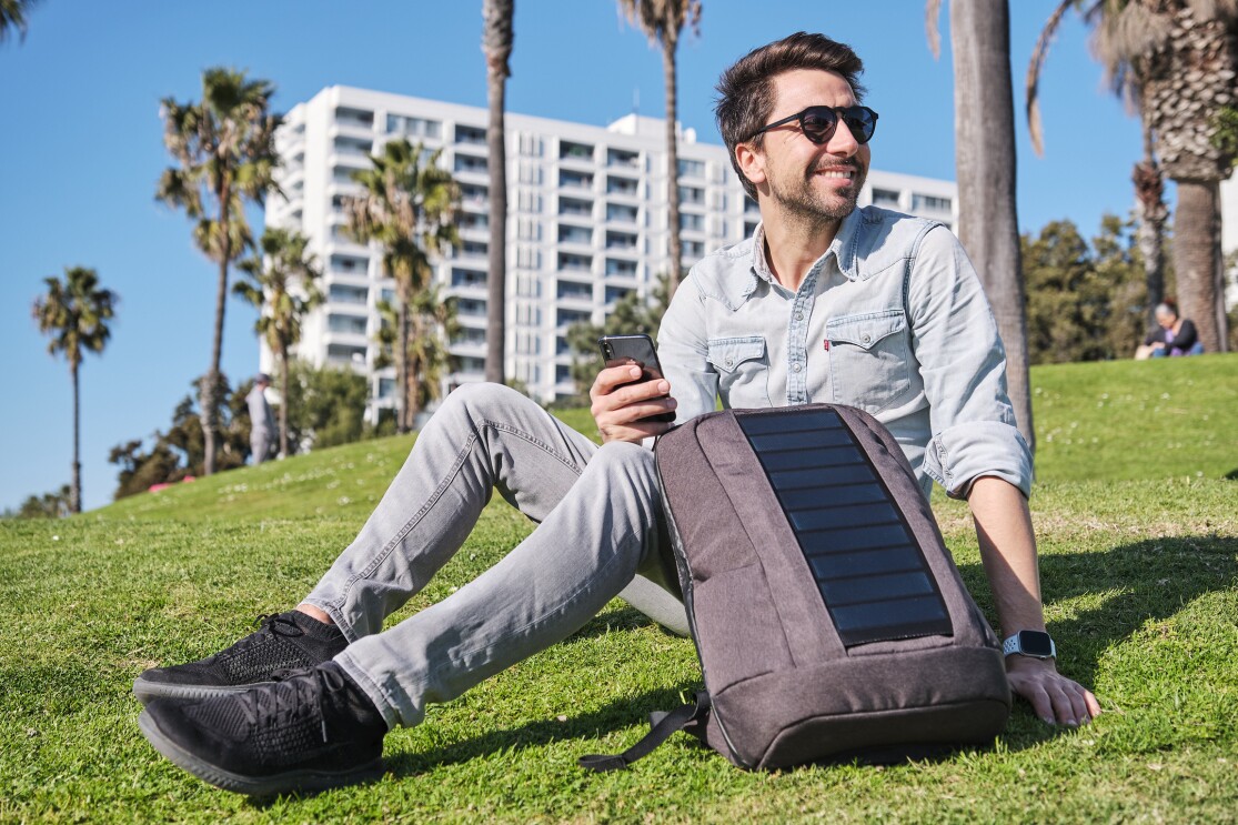 Ein Mann mit braunen Haaren und einem blauen Hemd sitzt auf einer Wiese. Neben sich hat er einen grauen Rucksack mit Solarpannels.