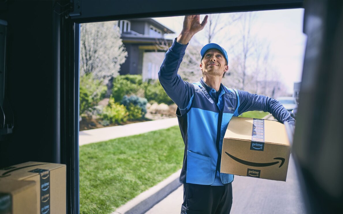 A photo of an Amazon delivery driver standing on the outside of their delivery van, holding a delivery package, closing the van door.