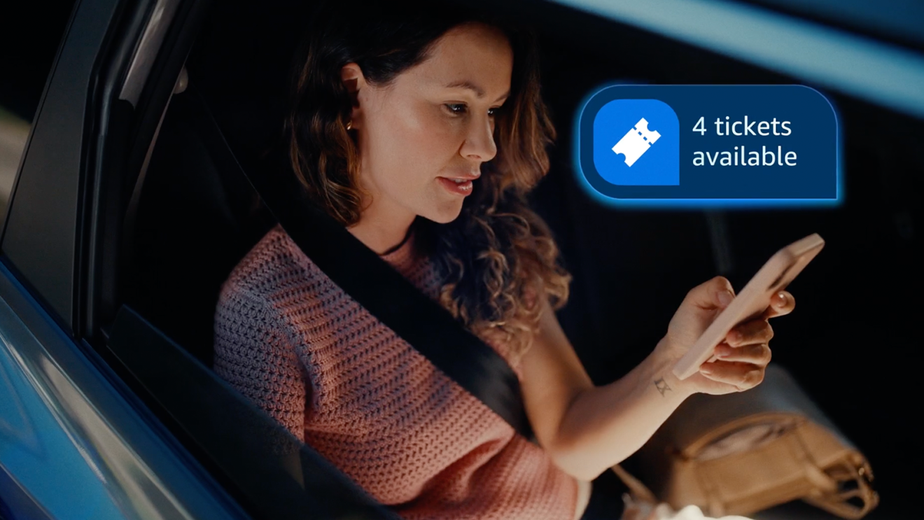 Woman in car checking phone, Alexa+ notification shows 4 tickets available