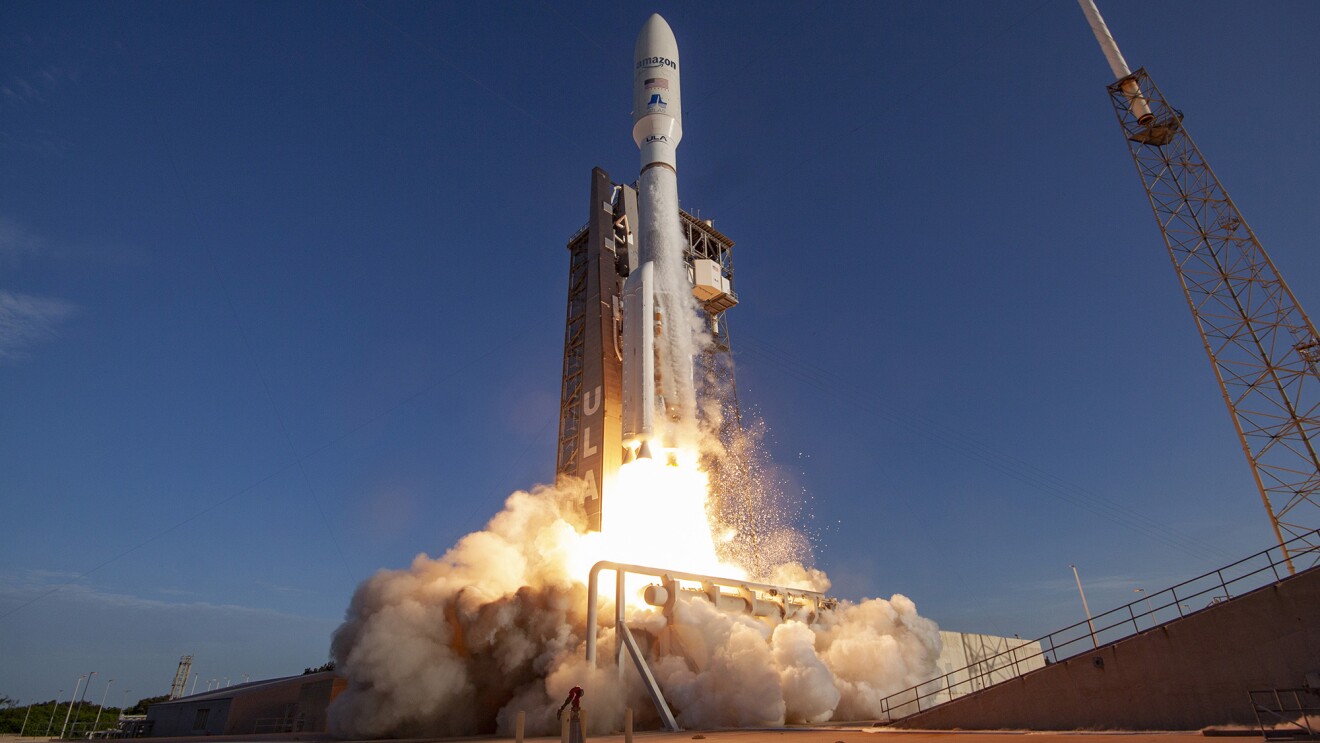 An image of a satellite launching. It has "Amazon" and the American flag decaled on the side. There is a blast below the satellite indicating launch and a blue, evening sky in the background.