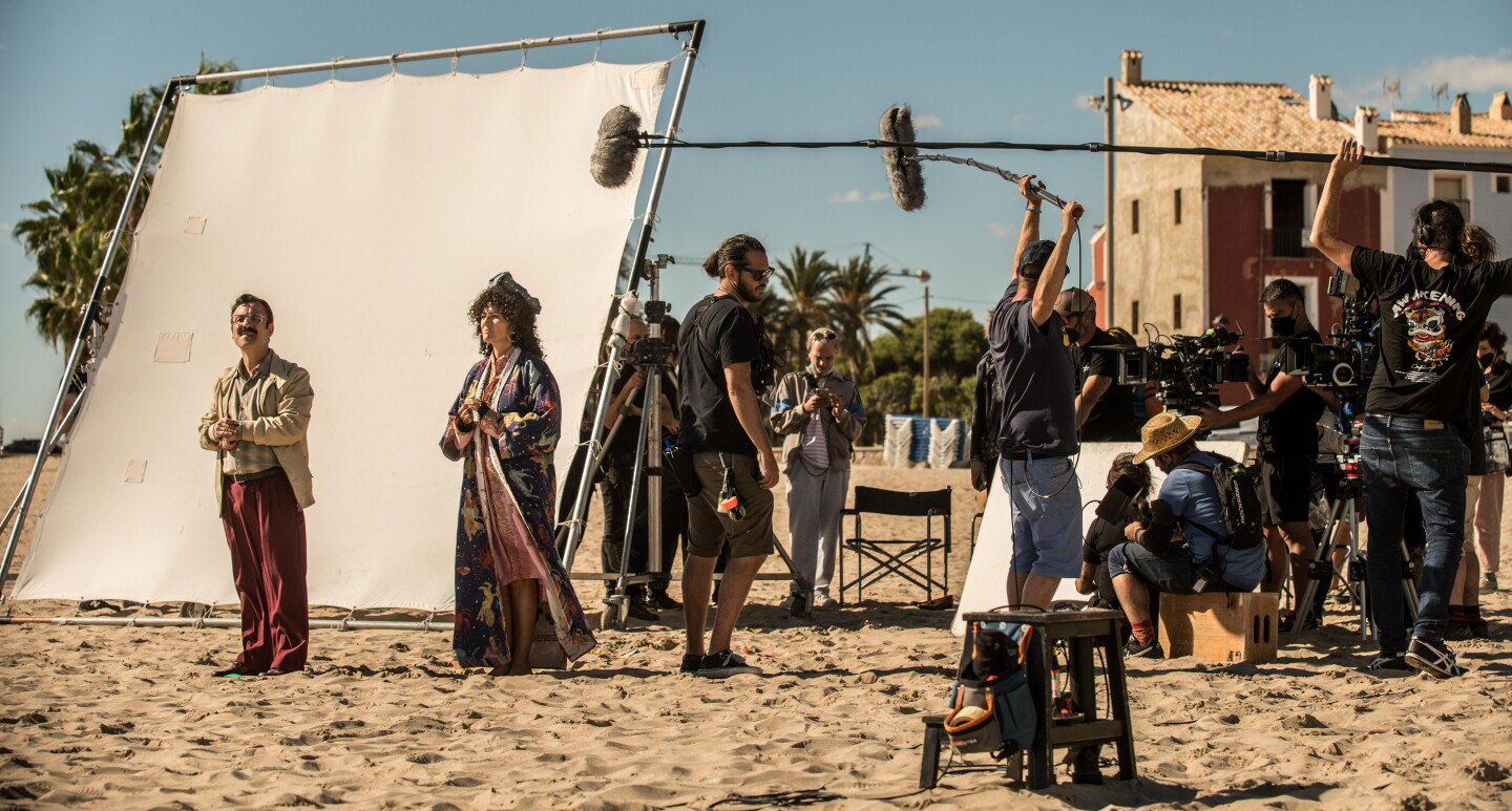 En un set de rodaje exterior, en la arena de la playa, aparecen dos actores, hombre y mujer vestidos de los años 60. Una pantalla blanca para aportiguar la luz. De fondo dos casa de pueblo marinero y, al lado de los actores, un equipo técnico de unas 10 personas con cámaras y micros. Todos van vestidos con ropa oscura. 