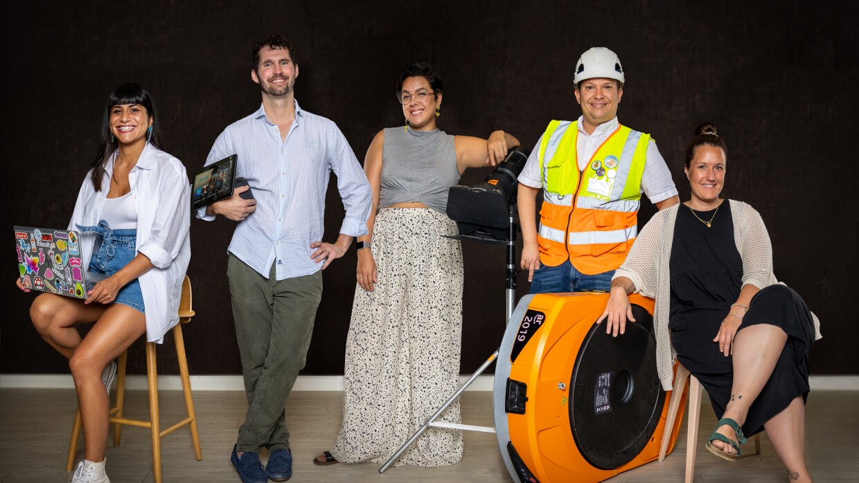 Es una foto de cinco trabajadores de Amazon posando en un tono informal. Hay tres mujeres y dos hombres. Las dos mujeres que están a la izquierda y a la derecha están sentadas y el resto de pie. Empezando de izquierda a derecha. Una mujer morena con el pelo largo, sentada en un taburete con su ordenador. Lleva una camisa blanca  con un jersey blanco debajo y unos pantalones cortos. Lleva una deportivas. El hombre de sui izquierda lleva el pelo corto y barba. Agarra una tablet. Lleva camisa azul por encima de un pantalón verde. La mujer que está en el centro está apoyada en un foto de cine de color negro. Lleva gafas, el pelo recogido, un jersey de color gris y una falda larga blanca con todos grises y negros. El hombre de su izquierda lleva casco y chaleco y está apoyado encima de un robot de Amazon. Debajo del chaleco lleva una camiseta blanca y unos jeans. La última mujer de la foto está sentada en una silla. Lleva el pelo recogida, apoya su brazo derecho en el robot. Lleva un vestido negro largo y por encima una chaqueta de punto. El fondo de la foto es negro.