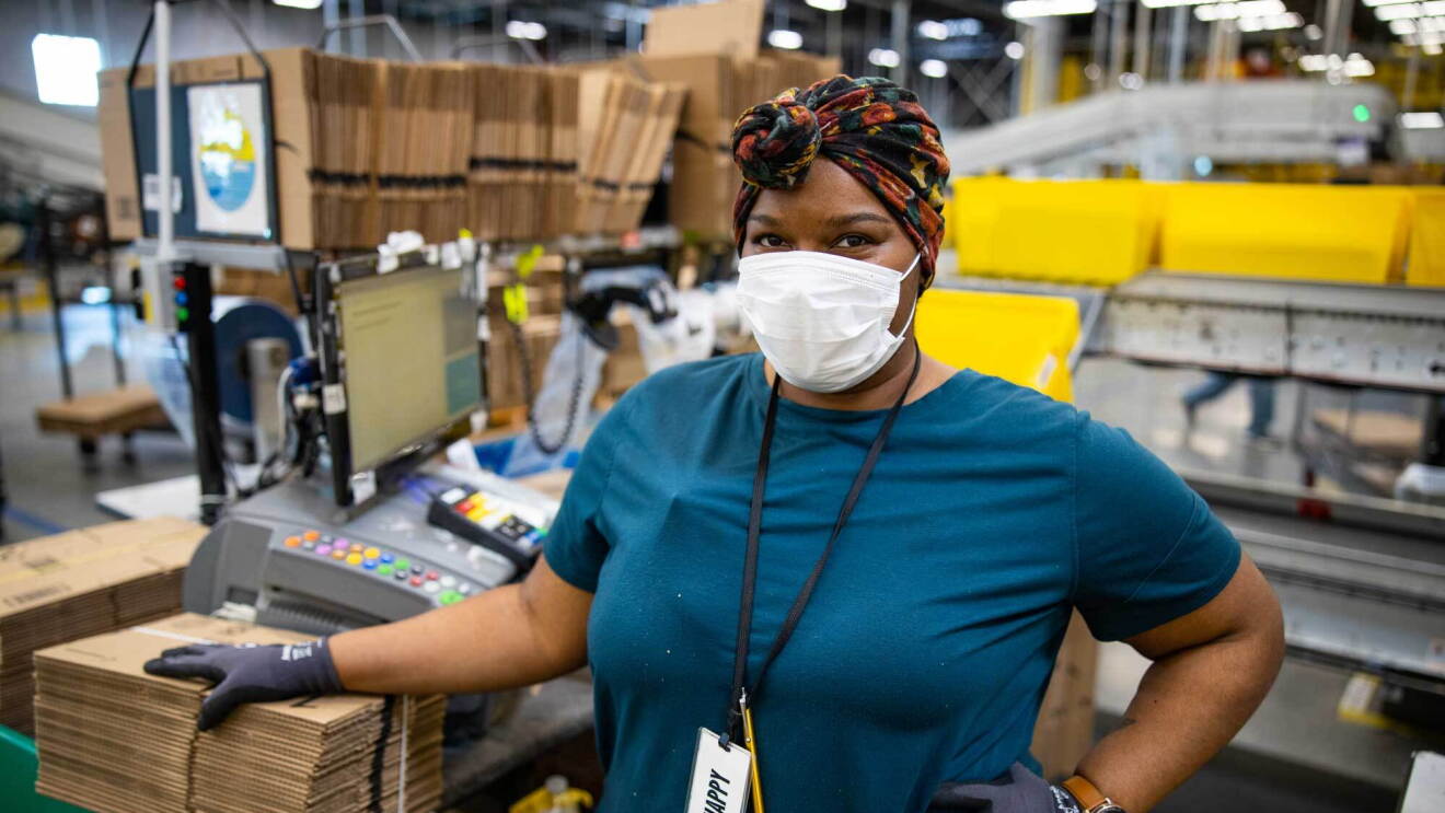 An Amazon associate working at a fulfillment center pack station poses for the camera while wearing a protective surgical mask