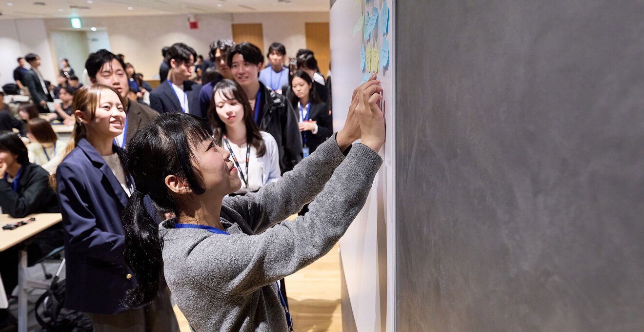 壁に貼られたホワイトボードに付箋を貼る若い女性の横顔。ボードにはすでにいくつか付箋が貼られている。奥に、順番待ちの列が伸びる。