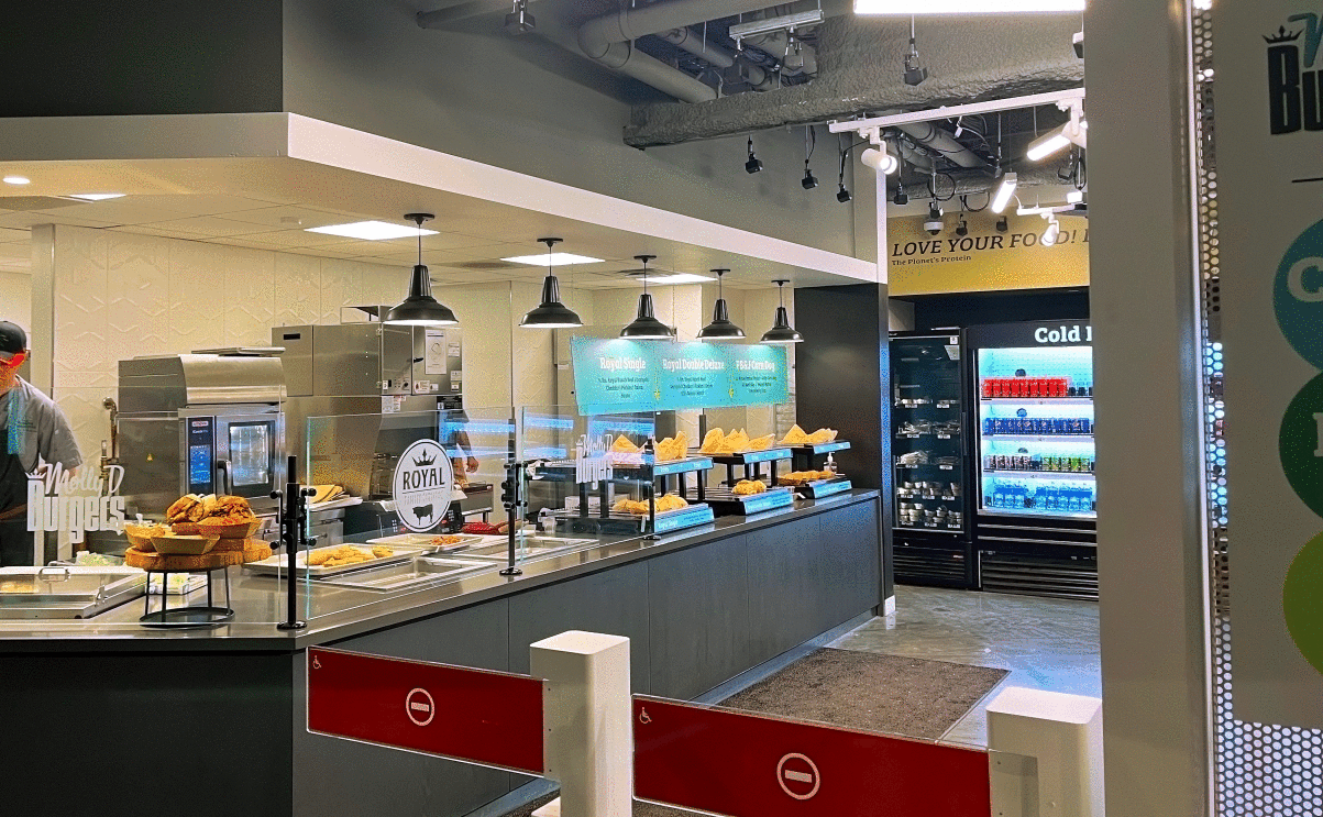 A counter of food with glass in front of it and people working in the background. There is a row of drink options in front of the glass where customers can line up to get food.