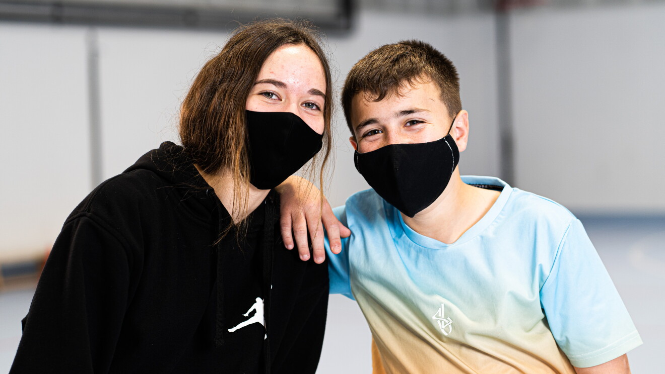Alumnos de 2 de ESO del IESO El Melgar  (Yuncos, Toledo). Los dos vam con mascarillas negras y de gondo se ve unas pareces grises de una pista deportiva. La chica va con un jersey deportivo de color negro. Tiene el pelo largo y recogido con una cola. Y los ojos azules. El chico va con una camiseta azul y amarilla de manga corta. Apoya su brazo al hombro de la chica. Tiene el pelo corto y castaño. 