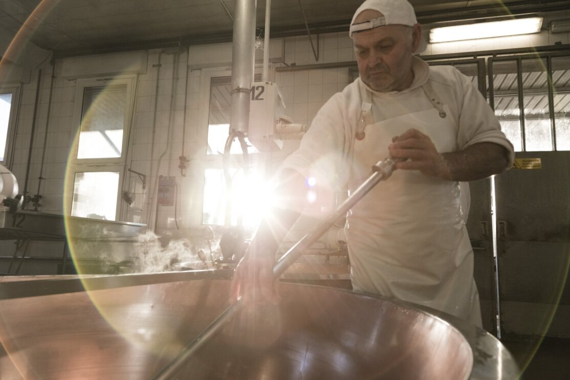 Immagine di un uomo mentre lavora il formaggio nel laboratorio della Latteria Due Madonne. L'uomo regge lo strumento chiamato 'spino' e con questo gira la cagliata dentro un grosso pentolone. L'uomo è vestito di bianco e indossa un grembiule e un cappello. La scena è ripresa di giorno. 