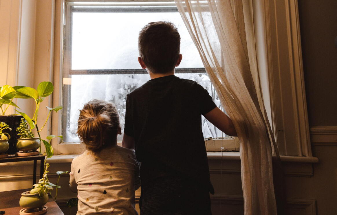 Doa heramos: una hermana de unos 3 años y un hermano de unos 7 años. Los dos están de espaldas y mirando por una ventana. El niño va con una camiseta negra de manga corta. La niña, con una camiseta blanca y con lágrimas de colores. Fuera de la ventana se ve todo blanco y parece que hay un árbol con nieve. El niño con la mano derecha aguanta unas cortinas blancas. El marco d e la ventana es blanco y de madera. En laparte izquierda de la foto hay un mueble con 4 plantes y se nota que entra la luz de una lámpara. 