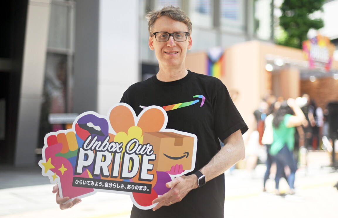 胸にレインボーカラーのAmazonのロゴが入っている黒いTシャツを着た男性。手にはUnbox Our PRIDEと書かれたパネルを持っている。後ろにビルとブースが見える