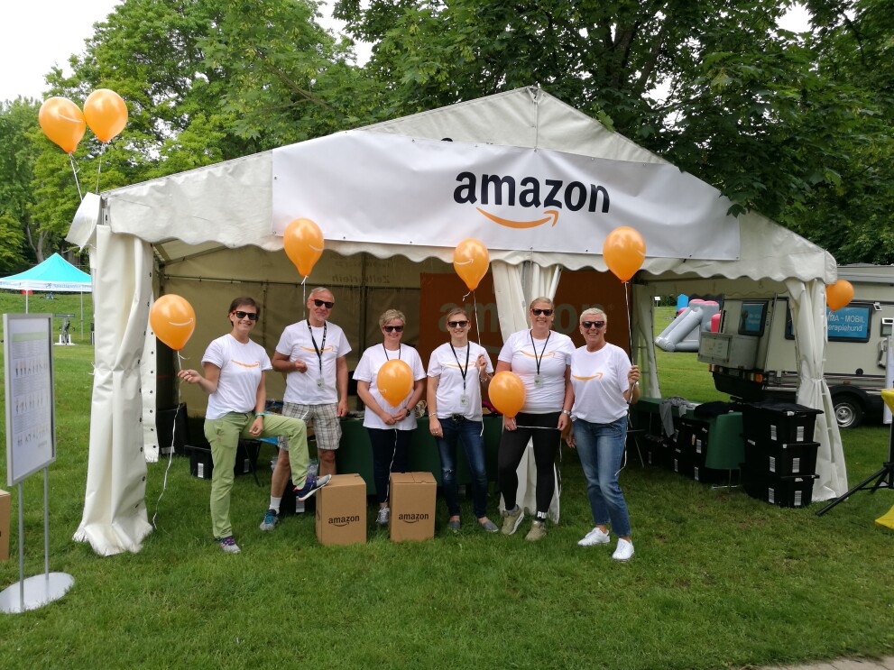 Eine Gruppe von Mitarbeitern in Amazon T-Shirts steht vor einem Zelt mit dem Amazon Logo. Einige tragen orange-farbene Luftballons.