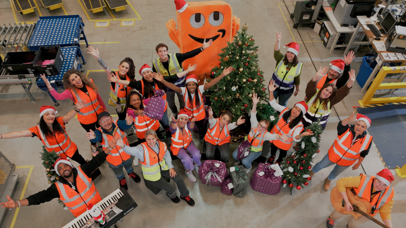 Gruppenbild mit Amazon Mitarbeiterinnen und Mitarbeitern aus Graben, alle tragen Weihnachtsmützen und orangene Sicherheitswesten. In der Mitte steht ein Weihnachtsbaum und um ihn herum Geschenke.