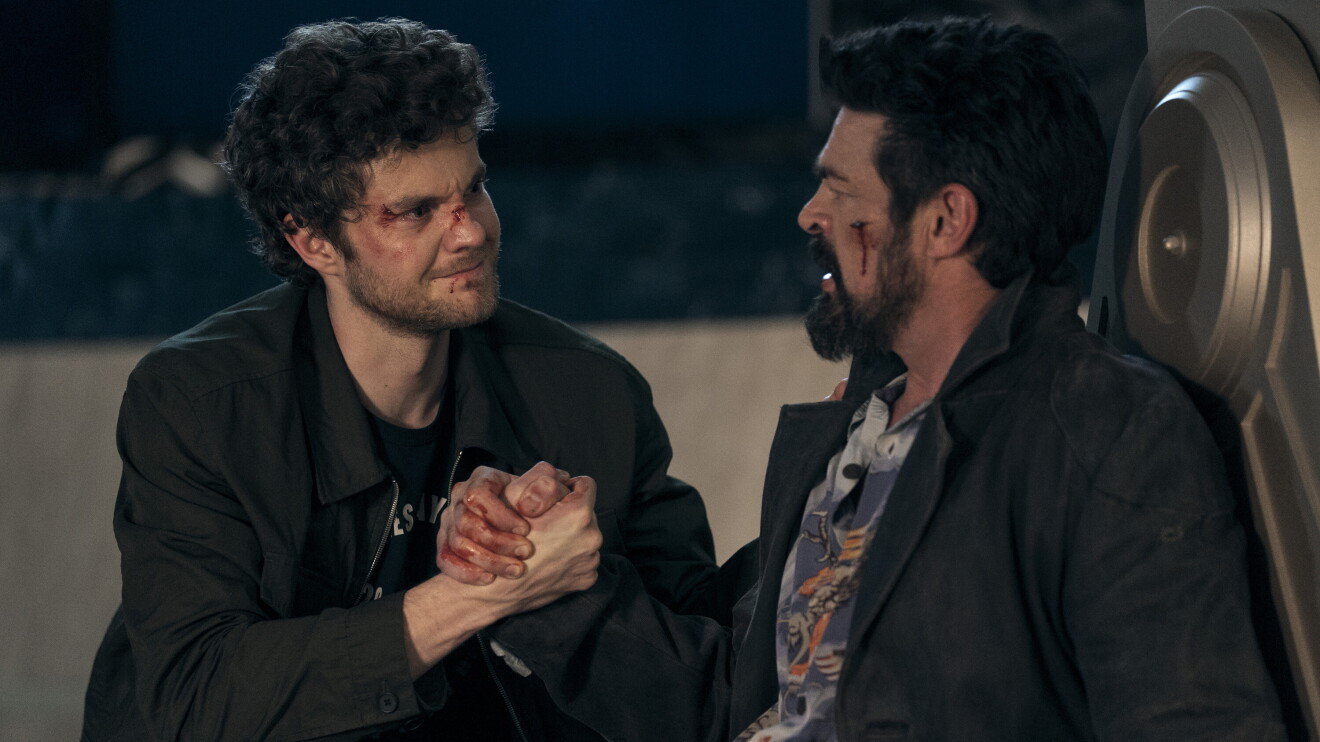 Two men in dark jackets clasping hands in a tense confrontation, one with visible injuries on his face