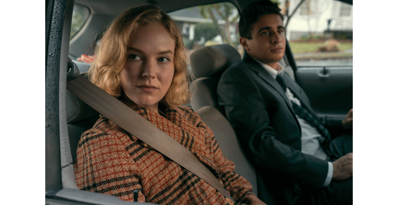 車の座席に並んで座る、チェック柄のコートを着た金髪の女性と、ダークスーツを着た黒髪の男性。女性だけシートベルトをしている。