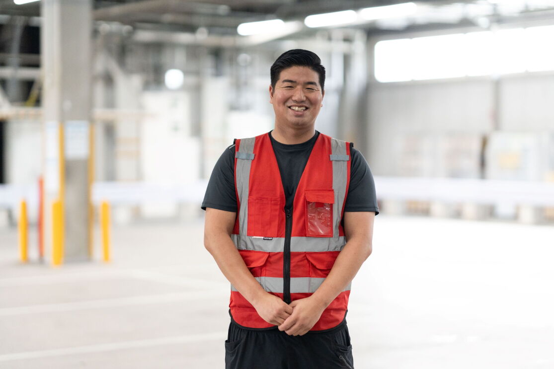 広い倉庫のような空間で黒いTシャツと赤い安全ベストを着ている男性が立っている。