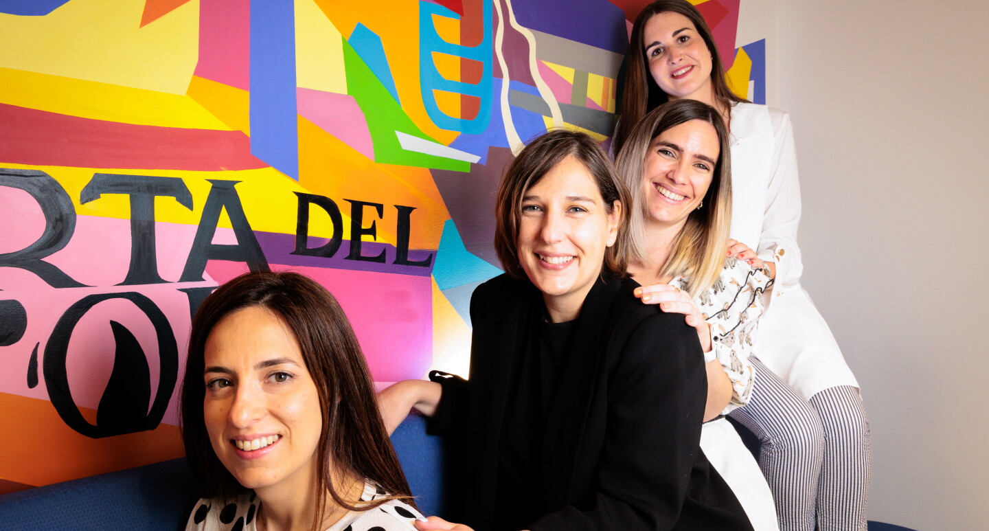 Sentadas en un sofá de color azul y con un fondo pintado de muchos colores vivos y las letras en negro de Puerta del Sol, están sentadas cuatro mujeres con la pierna izquierda cruzada. De izquierda a derecha: María Alonso vestida con un traje pantalón de color negro, con un jersey negro también debajo de la americana. Lleva unas sandalias marrones con dos tirantes. Al lado está Alejandra Ossa tiene el pelo negro largo hasta el hombro. Lleva un vestido blanco de lunares negros de manga larga y largo hasta la rodilla.  Lleva unas sandalias de color de oro de plata. Lara Fajardo es la cuarta de la foto. Va con una camisa blanca estampada, con el pelo hasta el hombro con mechas negras y decoloradas hasta el rubio en las puntas. Con pantalones blancos y unas sandalias negras de plataforma de color de mimbre de color marrón y negro. Y, finalmente, Miriam Domínguez-Adame Aguilera con el pelo liso negro hasta el pecho. Unas sandalias negras, unos pantalones rayados de color negro y blanco, una camisa y chaleco de color blanco.