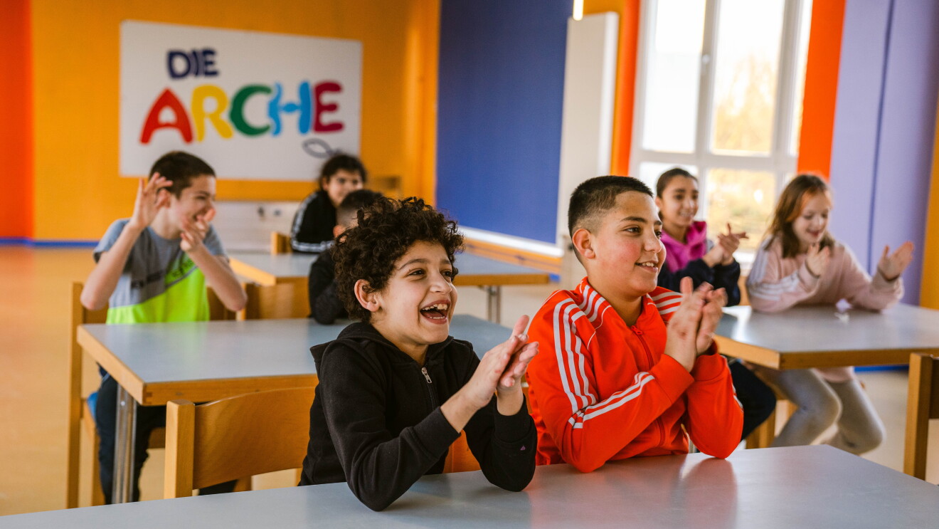 Kinder sitzen in einem Klassenzimmer, jeweils zu zweit an einem Tisch, sie schauen nach vorne und haben Spaß, lachen, klatschen in die Hände. Im Hintergrund hängt das Logo der Organisation "Die Arche".