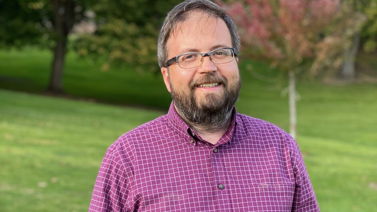 A man smiles at the camera. He wears glasses and a button down, and stands in a park setting.