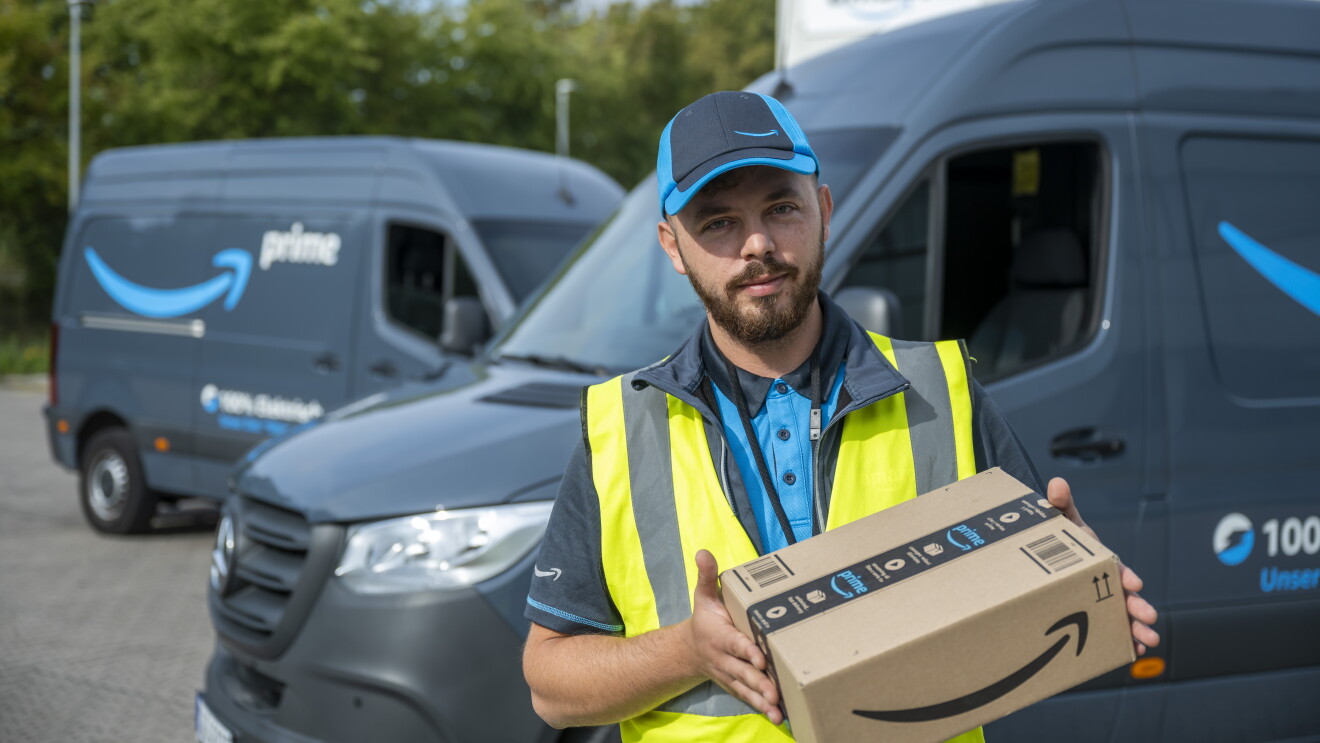 Ein Amazon Lieferfahrer mit einem Paket steht vor einem blauen Lieferfahrzeug.