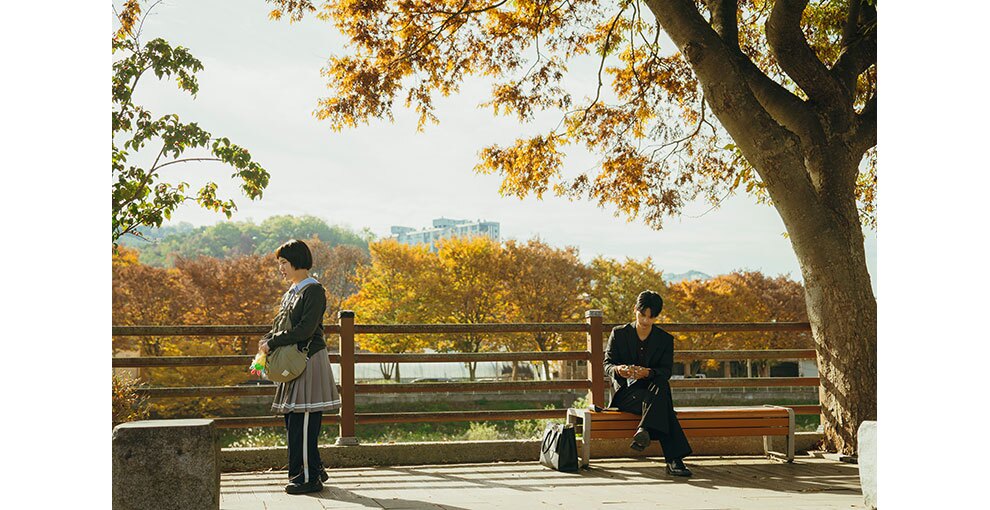 明るい屋外、公園のような場所。ベンチに座った男性と、その近くに男性に背を向けて立つ少女。木々の葉は黄色く色づいている。