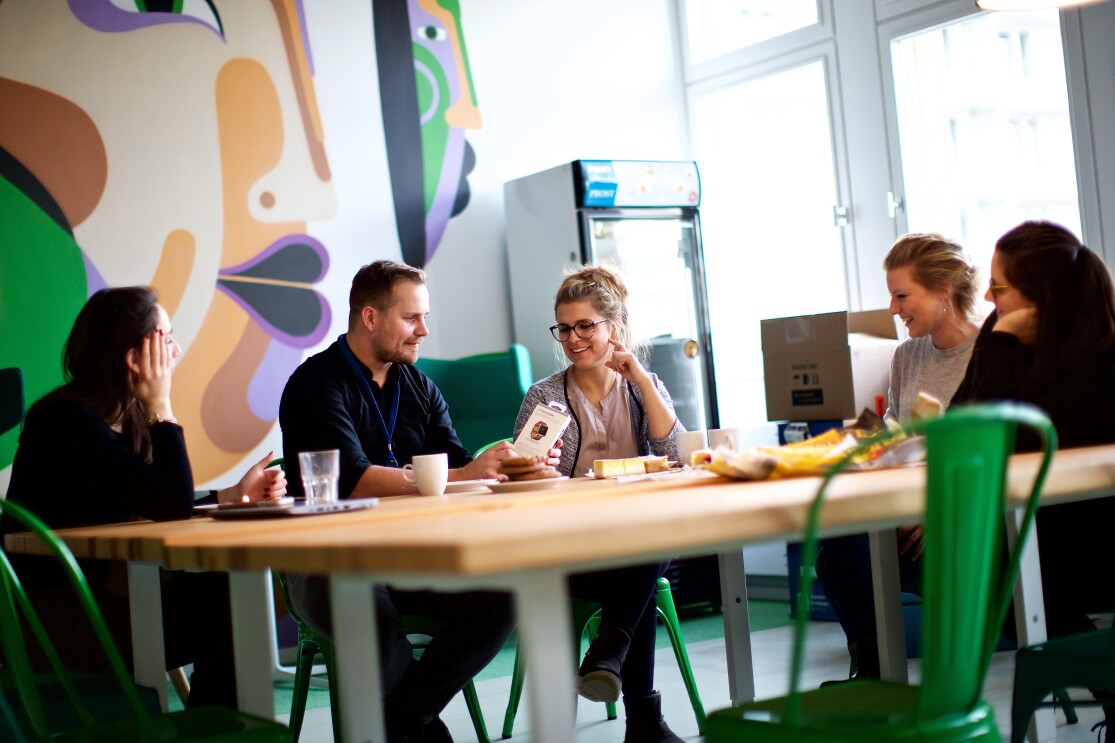Mitarbeiter sitzen im Bür an einem großen Gemeinschaftstisch, unterhalten sich, trinken Kaffee. Die Umgebung ist bunt gestaltet mit grünen Stühlen und farbigen Wänden.
