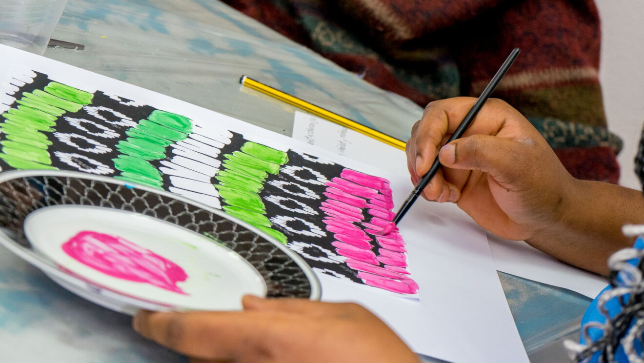 Primer plano de la las manos de una persona racializada pintando con un pincel sobre un papel. En la mano derecha tiene un pincel y en la izquierda agarra un plato con pintura de color rosa. En el caballete donde está pintando, que es inclinado, tiene también un papel con apuntes y un lápiz. 
