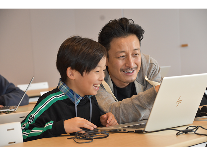 長机に載せたノートパソコンの画面を一緒に見つめる子供と男性。子供は笑顔を見せている。