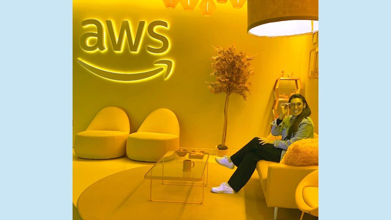 Amazon employee Annie sits in an AWS office lounge while touching her eyeglasses and sitting on a couch. 