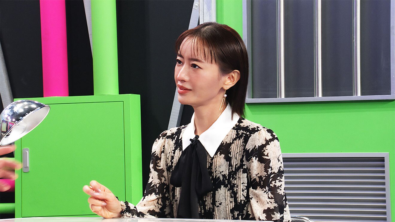 壁やパイプが派手な黄緑とピンクに塗られたセットで座る女性の上半身。胸元に黒いリボンのある黒字に白い花模様のワンピースで、片手を机の上にのせている。左側に手だけ写りこんでいる相手を見ている。