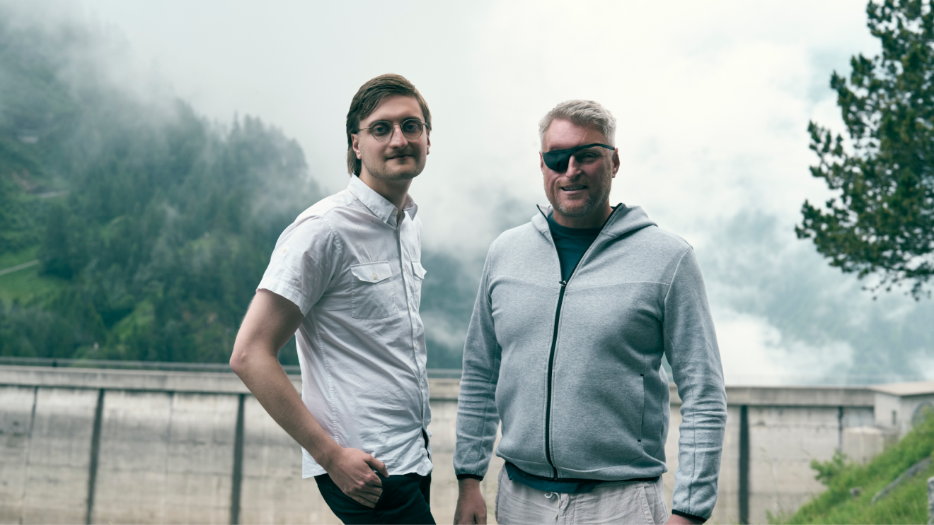 A young man and an older man wearing an eyepatch standing in front of a dam and mountains