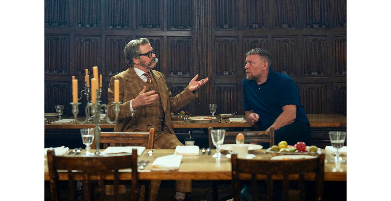 装飾的な羽目板壁の食堂で、三つ揃いスーツの男性とポロシャツの男性がテーブルにもたれて話している。並んだテーブルには燭台、グラス、皿が置かれている。