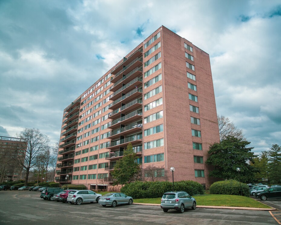 An image of a large apartment building. It has a parking lot with cars in front of it, green grass and tress to either side, and a blue sky with white clouds in the background.