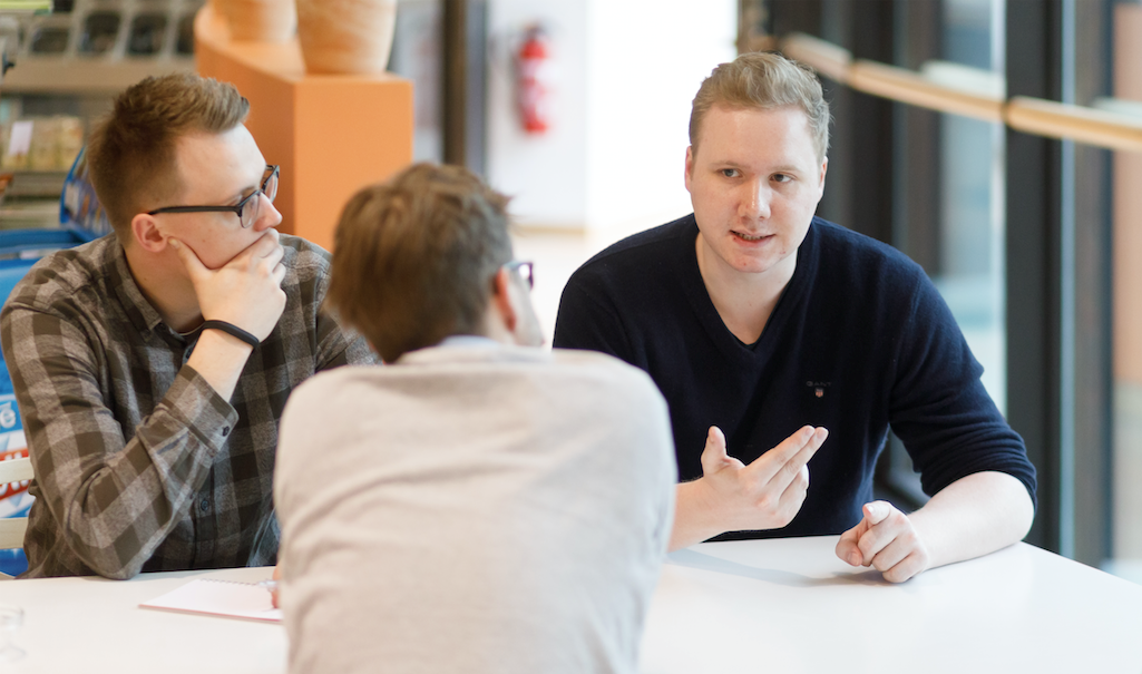 Drei junge Männer sitzen am Tisch und überlegen (zwei sieht man von vorne, einer hat den Rücken zur Kamera gewandt)