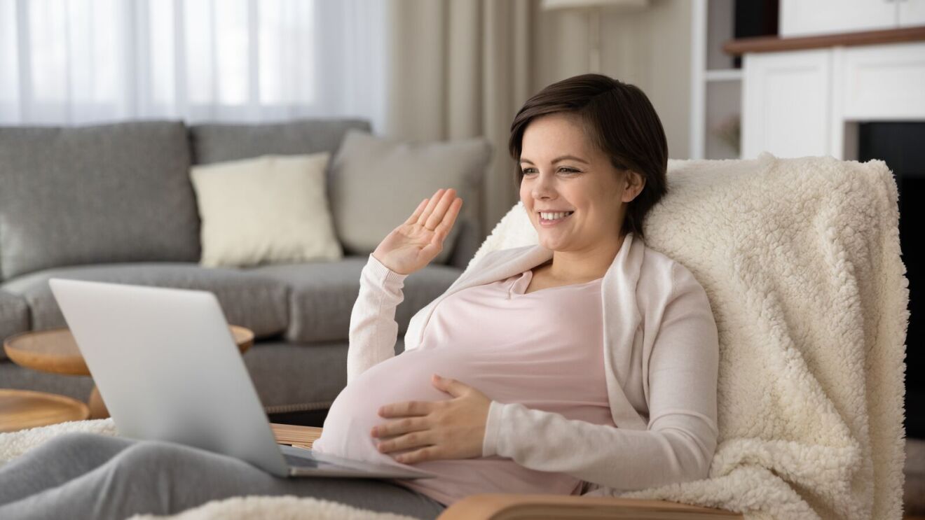 Pregnant woman waving at laptop during video call at home