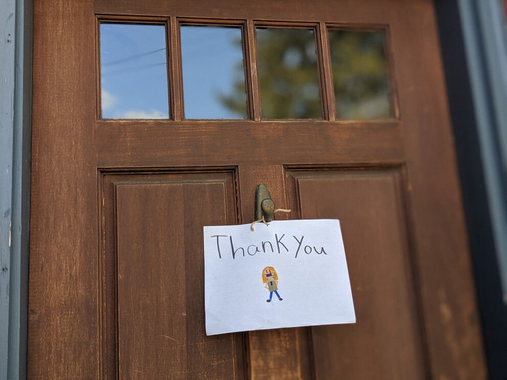A sign attached to the front door of a home, with a delivery driver wearing a mask, and "Thank you" drawn on it.