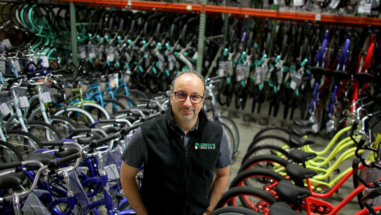 一名男子微笑着在自行车店拍照,身后是一排排的自行车。他穿着一件背心,上面写着“孔蒂自行车店”。