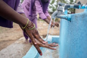 A photo of two people washing their hands at an outdoor faucet. 