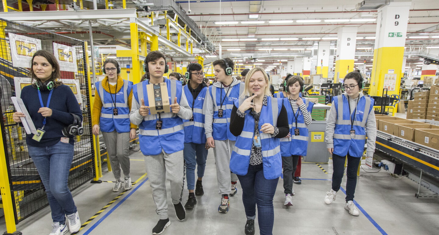 Una foto di Daria Banach, Tour Lead nel centro di distribuzione Amazon di Passo Corese, mentre accompagna i ragazzi di una scolaresca in un tour del magazzino. Daria parla al microfono e i ragazzi sono muniti di cuffie.
