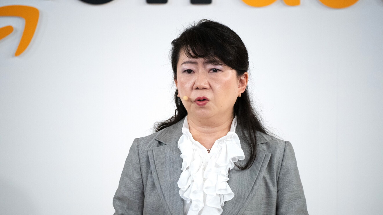 フリルのついた白いブラウスとグレーのジャケットを着た女性がヘッドセットをつけて話している（二村真理子氏）