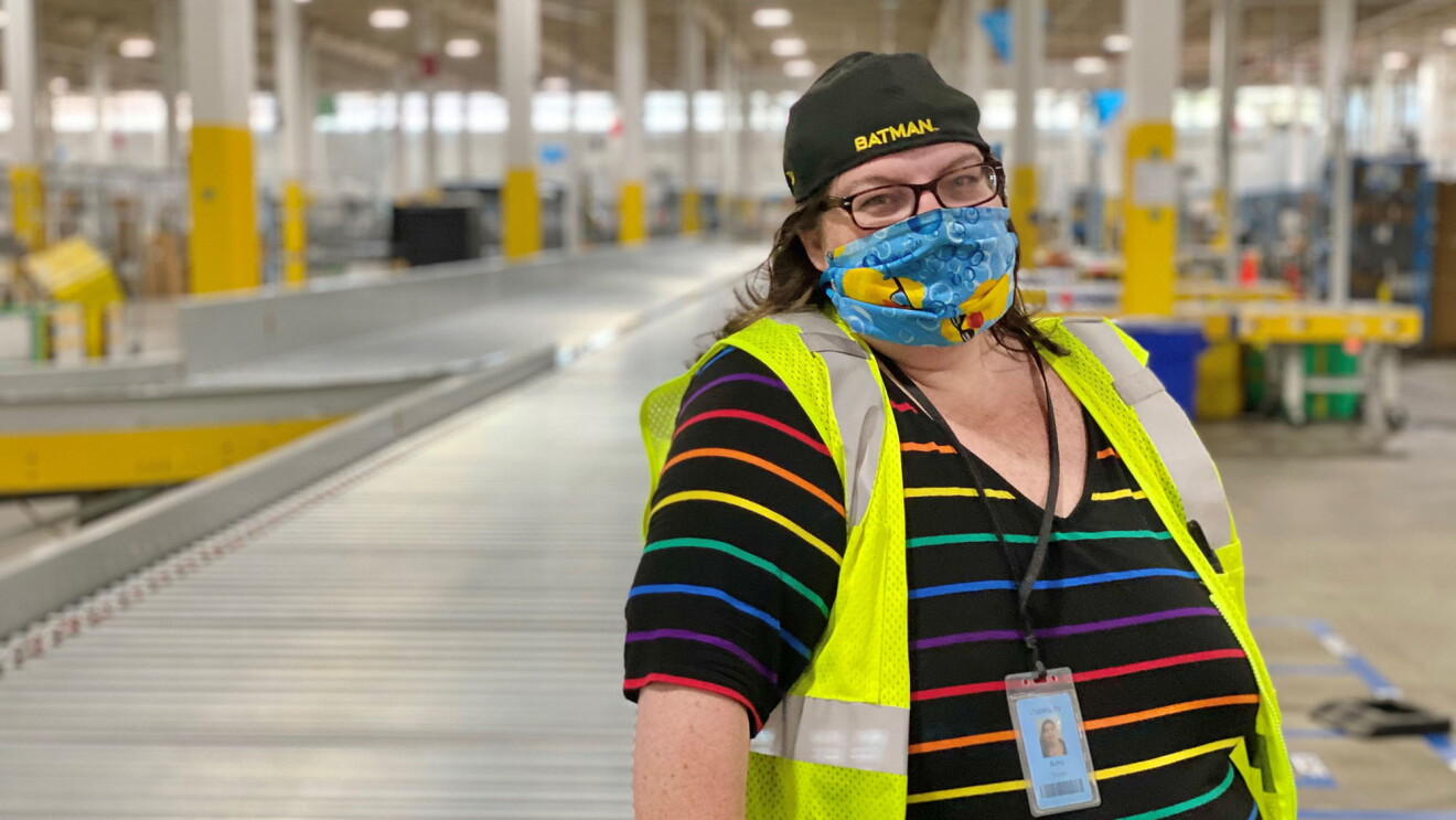 A woman wearing a rainbow-striped shirt, a colorful face mask, and a yellow safety vest stands in an Amazon fulfillment center, smiling at the camera from behind her mask. 