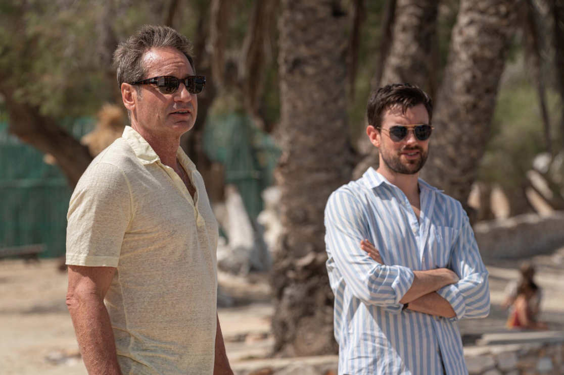 Two men (David Duchovny and Jack Whitehall) stand in the sun wearing sunglasses and looking into the distance