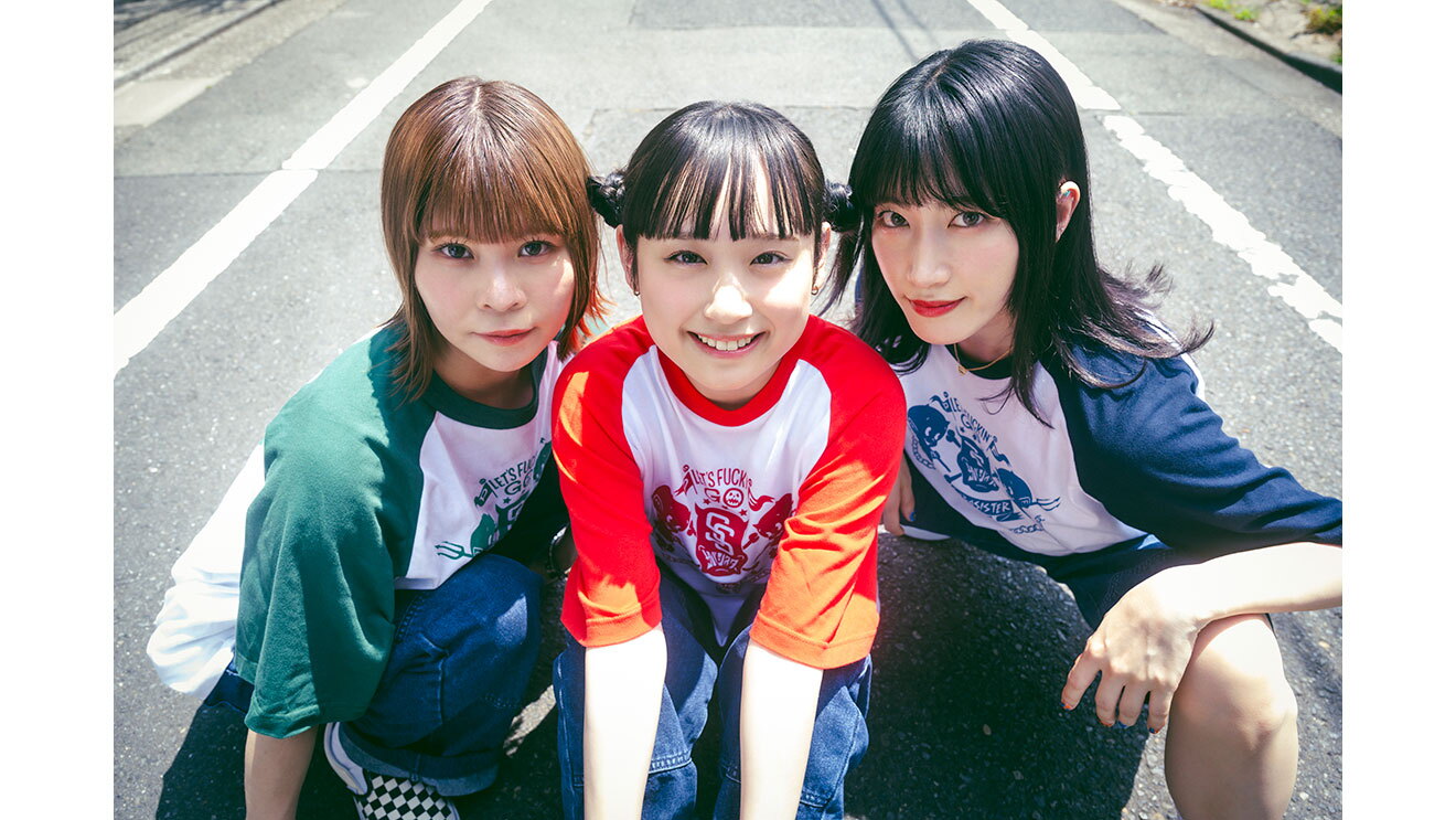 3人の女性が道路上でしゃがみ、笑顔でこちらを見ている。色違いのお揃いTシャツを着ている。