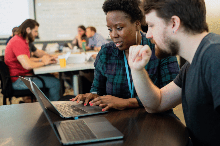A white male employee follows along as a Black female employee walks him through work on her laptop.