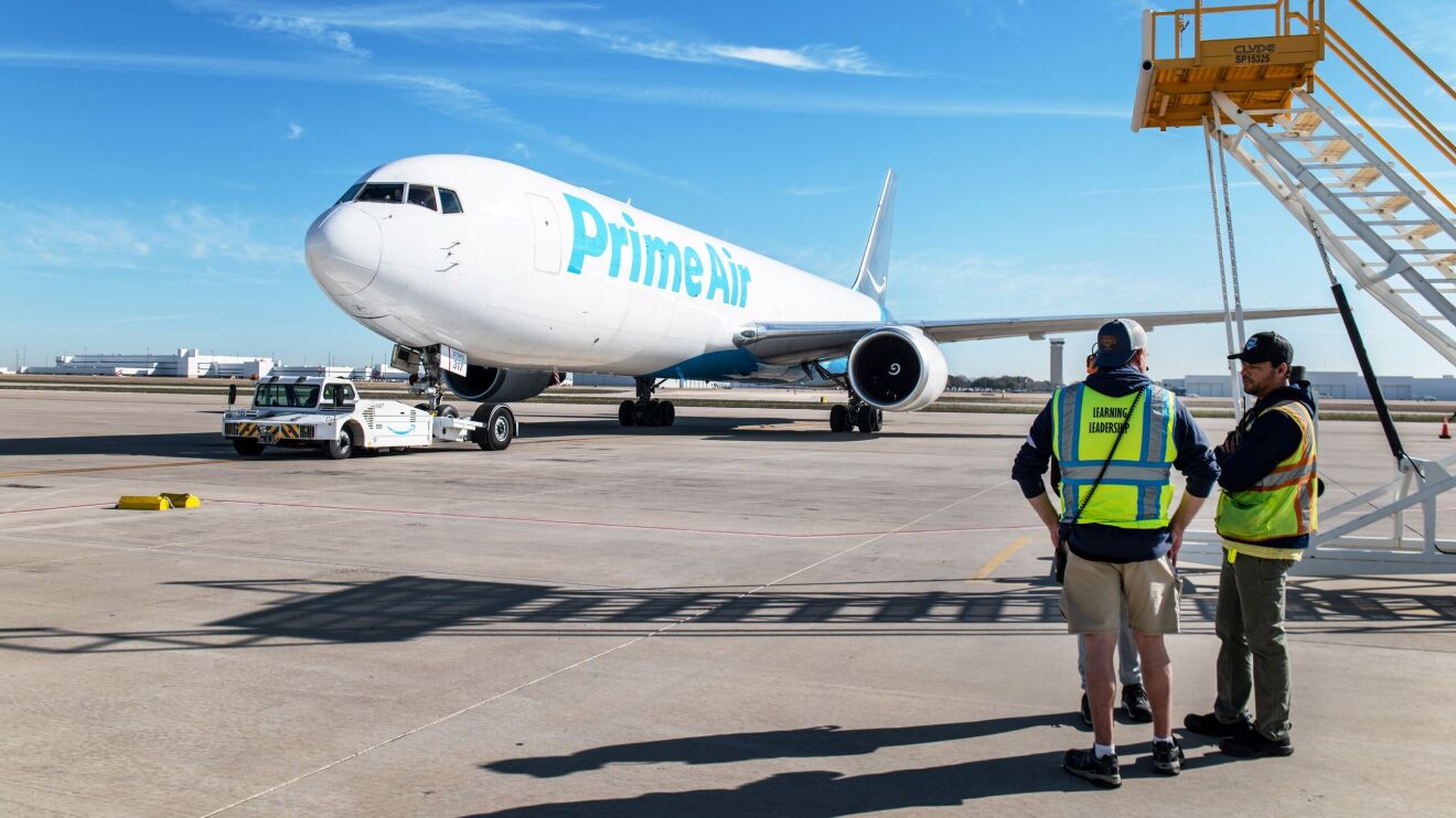 An image of an Amazon Air plane parked outside on an air hub. There is a small car pushing the plane back onto the taxiway.
