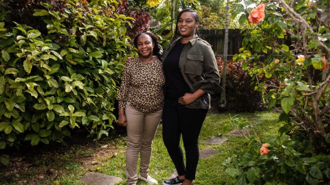 Kimberly and her mom stand next to one another with their arms around each other. They stand in a beautiful, green, luscious backyard. Kimberly is wearing black pants with a black shirt and an olive green jacket. Her mom wears khaki pants and a leopard top.