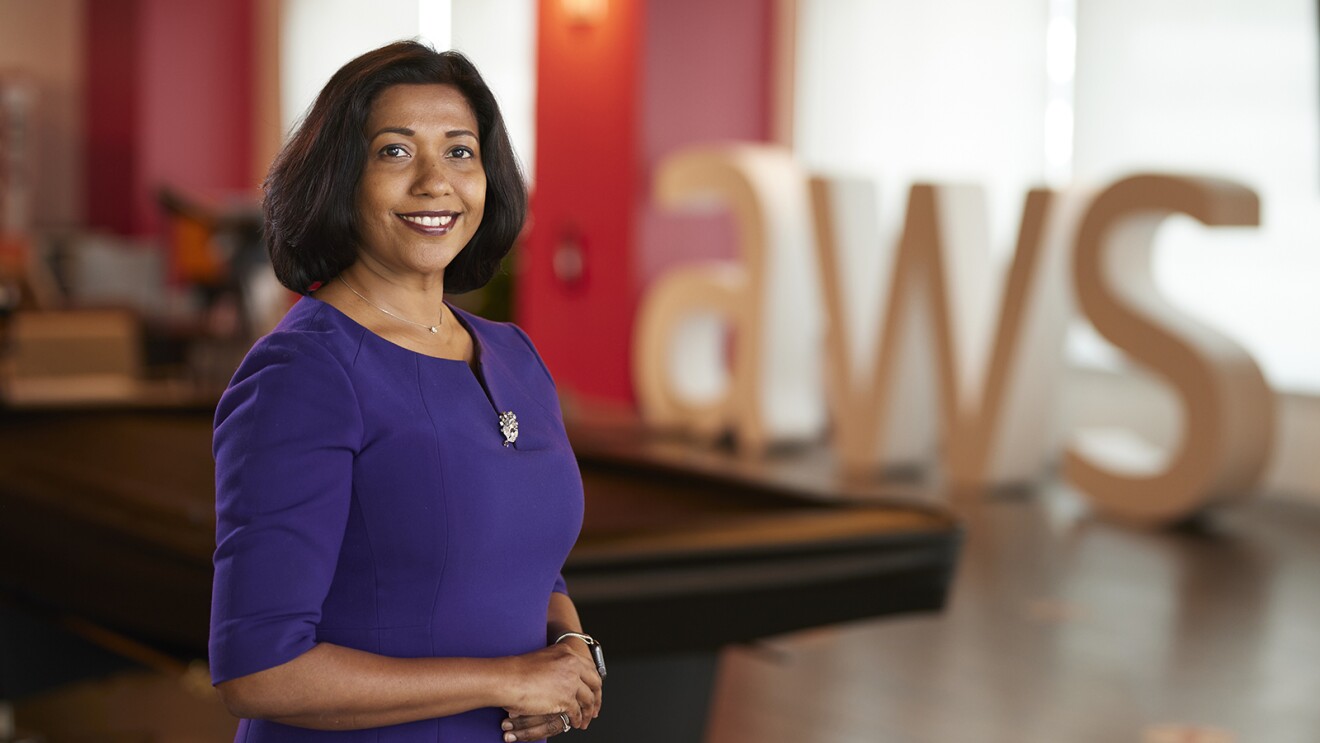 An image of a woman standing and smiling for a photo in a game room at an Amazon office. There is a large, letter sign in the background that says "AWS."