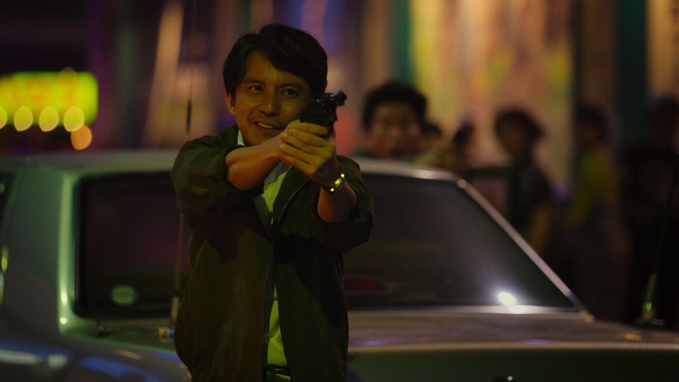 薄暗い場所で車の前に立ち、両手で拳銃を構える若い男性